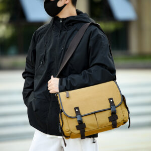 A stylish man is carrying a vintage olive green canvas messenger bag across his shoulder in an urban environment, showcasing his classic daily commuting look.